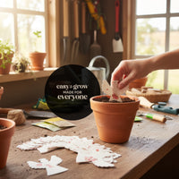 A hand placing a white plantable seed paper angel funeral favor decorated with pressed pink flowers into a terra cotta pot filled with soil