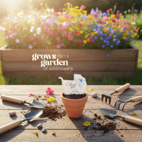 Gardening tools and a pot with a baby carriage seed paper baby shower favor by Seed Bloom gifts on top of it with wildflower seeds on a wooden table, with a garden bed of flowers in the background.