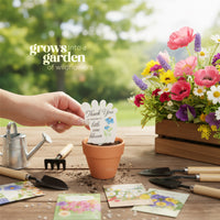 Hand placing a
Baby footprint seed paper baby shower favor by Seed Bloom Gifts, into a pot with a garden of wildflowers in the background.