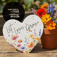 Heart-shaped planter with 'Let Love Grow' text and embedded wildflower seeds, next to a pot of flowers.