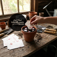 A hand lifts a flower pot containing a "Bloom Thank You" card, showcasing a thoughtful gesture