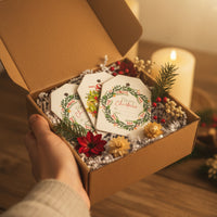 A hand holds an open, rustic cardboard box containing three Christmas Holiday Gift tags by Seed Bloom Gifts. Nestled in white shredded paper and decorated with pine sprigs, berries, and dried flowers,