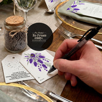 Seed paper gift tag with purple lavender botanical design being personalized with pen in elegant table setting, showing pre-printed 'To/From' fields for easy personalization and planting instructions, displayed with fine dining place setting and decorative elements