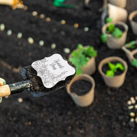 Overhead shot of a gardener planting the Floral Plantable Thank You Seed Paper Tags by Seed Bloom Gifts using a trowel in rich garden soil surrounded by peat pots.