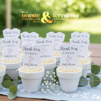 White pots with pink onesie plantable seed paper baby shower favors by Seed Bloom Gifts, featuring signs and baby-themed text, set against a blurred green outdoor background.