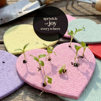Heart-shaped seed confetti with small plants on a wooden surface, with a circular text overlay.