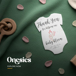 Plantable seed paper baby shower favor cut in a onesie shape, printed with 'Thank You for helping our baby bloom' text and a delicate pink branch illustration by Seed Bloom Gifts