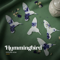 Two plantable seed paper favors cut in the shape of hummingbirds, featuring blue and purple watercolor-style pigmentation and embedded petals, shown packaged with a white planting instruction card by Seed Bloom Gifts