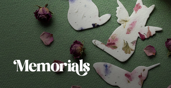 Plantable seed paper shapes (birds/doves and a hummingbird) with dried flowers embedded, next to dried rosebuds and petals, on a green background by Seed Bloom Gifts. The word "Memorials" is in the bottom left.