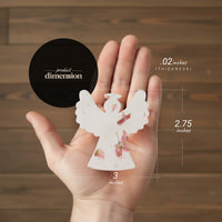 Hand holding a small angel-shaped object with dimensions indicated on a wooden background