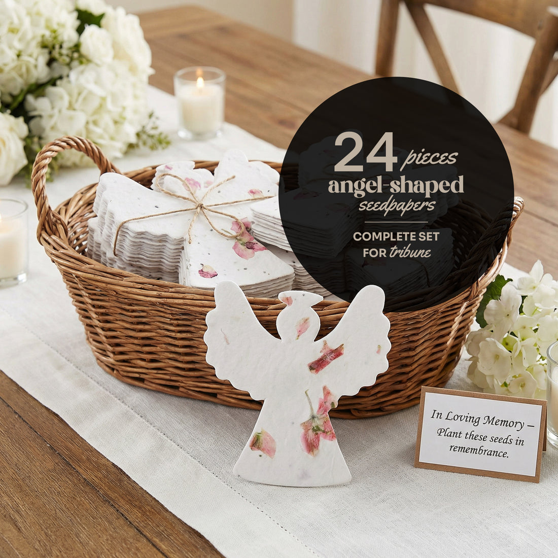 A basket filled with 24 white plantable seed paper angel-shaped funeral favors decorated with pressed flowers tied with twine, displayed with white flowers, a candle, and a sympathy card on a wooden table