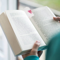 A person holding an open book near a window, using a tag that resembles a Pink Blank Tags Plantable Seed Paper by Seed Bloom Gifts with a dried pink petal as a bookmark.