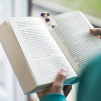 A person holds an open book near a window, using a tag that resembles a Purple Blank Tags Plantable Seed Paper by Seed Bloom Gifts, which contains a blue dot and embedded purple petals, as a bookmark.