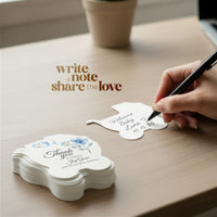 Hand writing on the back of a baby carriage seed paper baby shower favor by Seed Bloom Gifts with 'Write a note, share the love' text on a wooden surface.