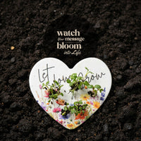 Heart-shaped white plate with plants growing on it, placed on dark soil with text 'Watch your message bloom into life'.