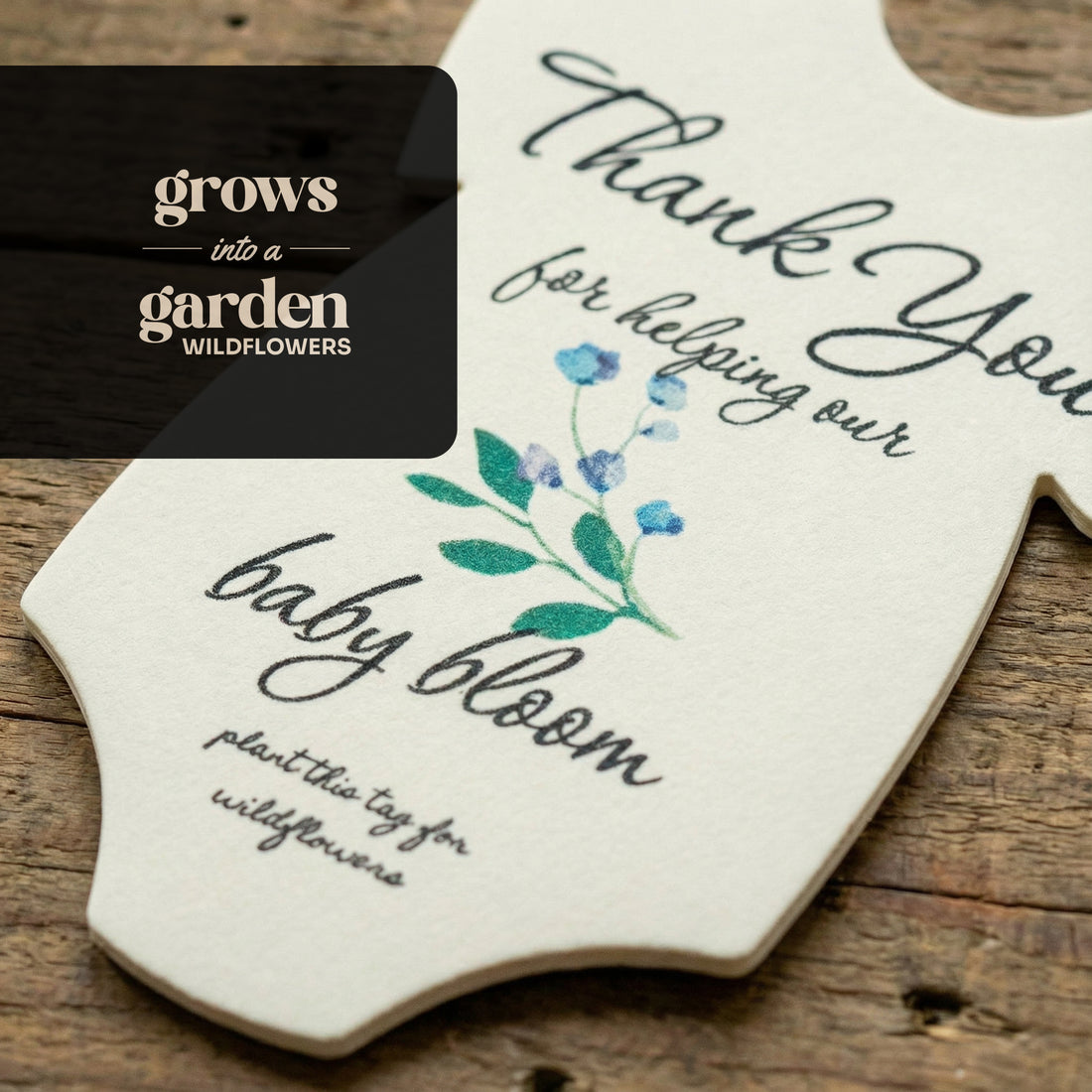 Close-up of a plantable seed paper onesie baby shower favor with blue floral illustration and the message “Thank you for helping our baby bloom,” shown on a rustic wooden surface with text noting it grows into wildflowers.