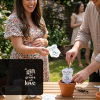 Pregnant woman and partner planting a plantable seed paper onesie baby shower favor into a terracotta pot at an outdoor gathering, smiling as the card with the message “Thank you for helping our baby bloom” is placed in soil.