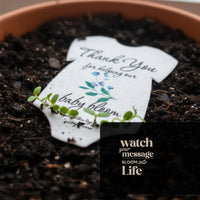 Close-up of a sprouting plantable seed paper onesie baby shower favor placed in soil, with small green seedlings emerging around the card that reads “Thank you for helping our baby bloom.”
