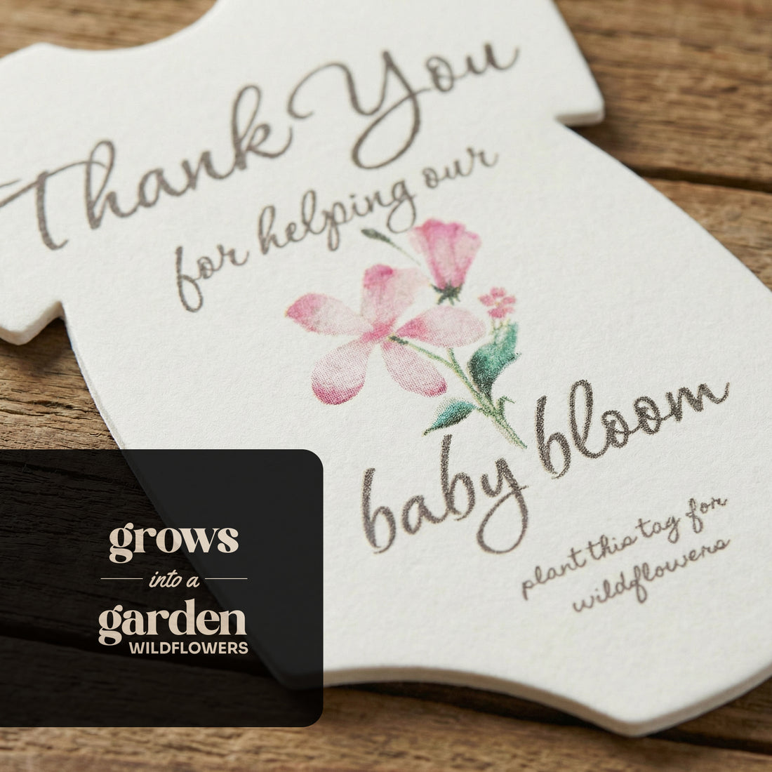 Plantable seed paper onesie baby shower favor by Seed Bloom Gifts, featuring a pink flower illustration and the text “Thank you for helping our baby bloom,” displayed on a rustic wooden surface.