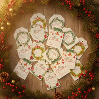 Overhead view of a variety of white Christmas Holiday Gift tags scattered on a wooden surface, surrounded by a festive pine and berry wreath. The tags feature four unique holiday designs, by Seed Bloom Gifts.