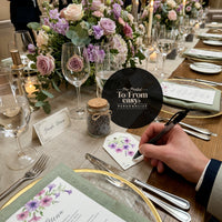 A man is seated at a table, writing next to a place card that reads "Bloom Thank You Tags