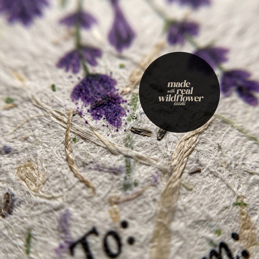 Close-up macro detail of seed paper gift tag showing embedded real wildflower seeds visible in cream cardstock texture with purple seed