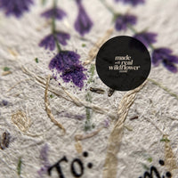 Close-up macro detail of seed paper gift tag showing embedded real wildflower seeds visible in cream cardstock texture with purple seed