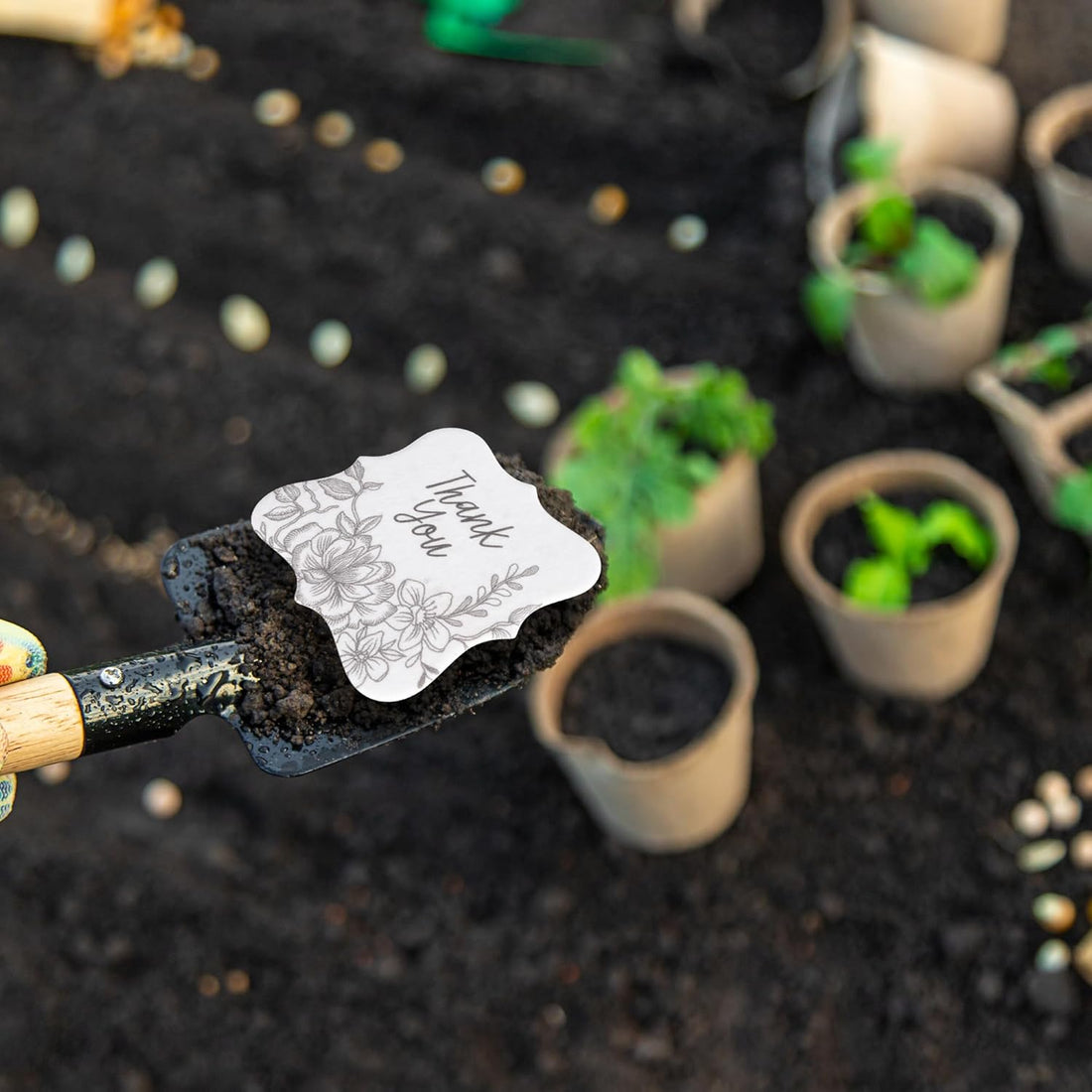 Overhead shot of a gardener planting the Floral Plantable Thank You Seed Paper Tags by Seed Bloom Gifts using a trowel in rich garden soil surrounded by peat pots.