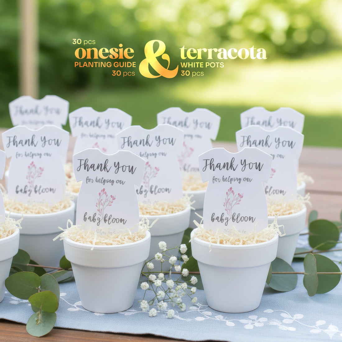 White pots with pink onesie plantable seed paper baby shower favors by Seed Bloom Gifts, featuring signs and baby-themed text, set against a blurred green outdoor background.