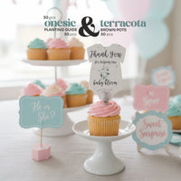The gender neutral green onesie plantable seed paper baby shower favor by Seed Bloom gifts is displayed on a pink cupcake stand next to pastel cupcakes and 'He or She?' and 'Sweet Surprise' party signs. 