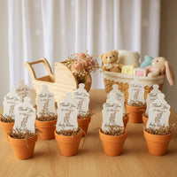 Small potted plants with 'Thank You' baby shower seed paper bundle by Seed Bloom Gifts  on a wooden surface, with a basket of toys in the background
