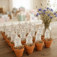 Small potted plants with 'Thank You' Baby Bottle Seed Paper favors by Seed Bloom Gifts on a wooden surface, with a vase of flowers in the background.