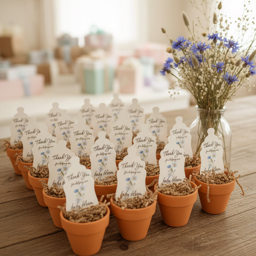 Small potted plants with 'Thank You' Baby Bottle Seed Paper favors by Seed Bloom Gifts on a wooden surface, with a vase of flowers in the background.