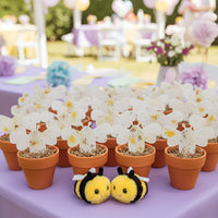 Decorative setup with Bee Shaped Seed Paper Baby Shower Favor By Seed Bloom Gifts in terracotta pots and plush bee toys on a purple tablecloth.