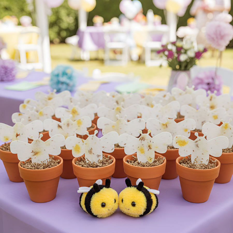 Decorative setup with Bee Shaped Seed Paper Baby Shower Favor By Seed Bloom Gifts in terracotta pots and plush bee toys on a purple tablecloth.