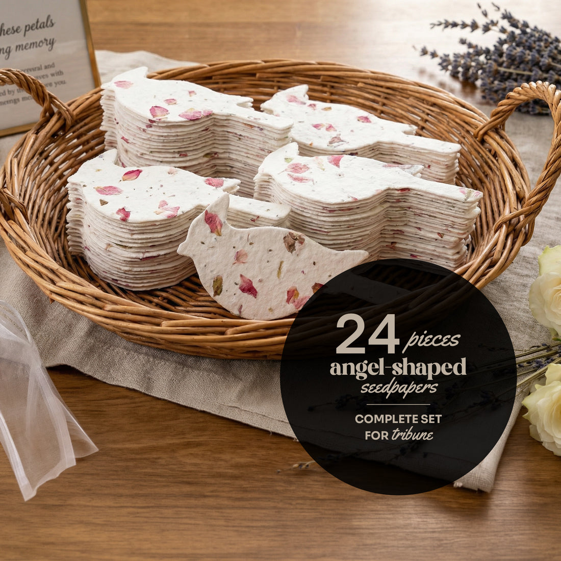 A basket filled with 24 white plantable seed paper angel-shaped funeral favors decorated with pressed pink flowers, displayed as a complete set for a tribute on a wooden table with lavender, white flowers, and a sympathy card