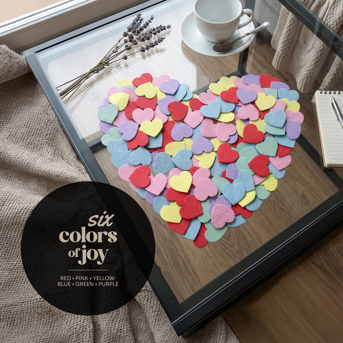 Heart-shaped arrangement of colorful heart-shaped confetti on a glass table with a cup and saucer in the background.