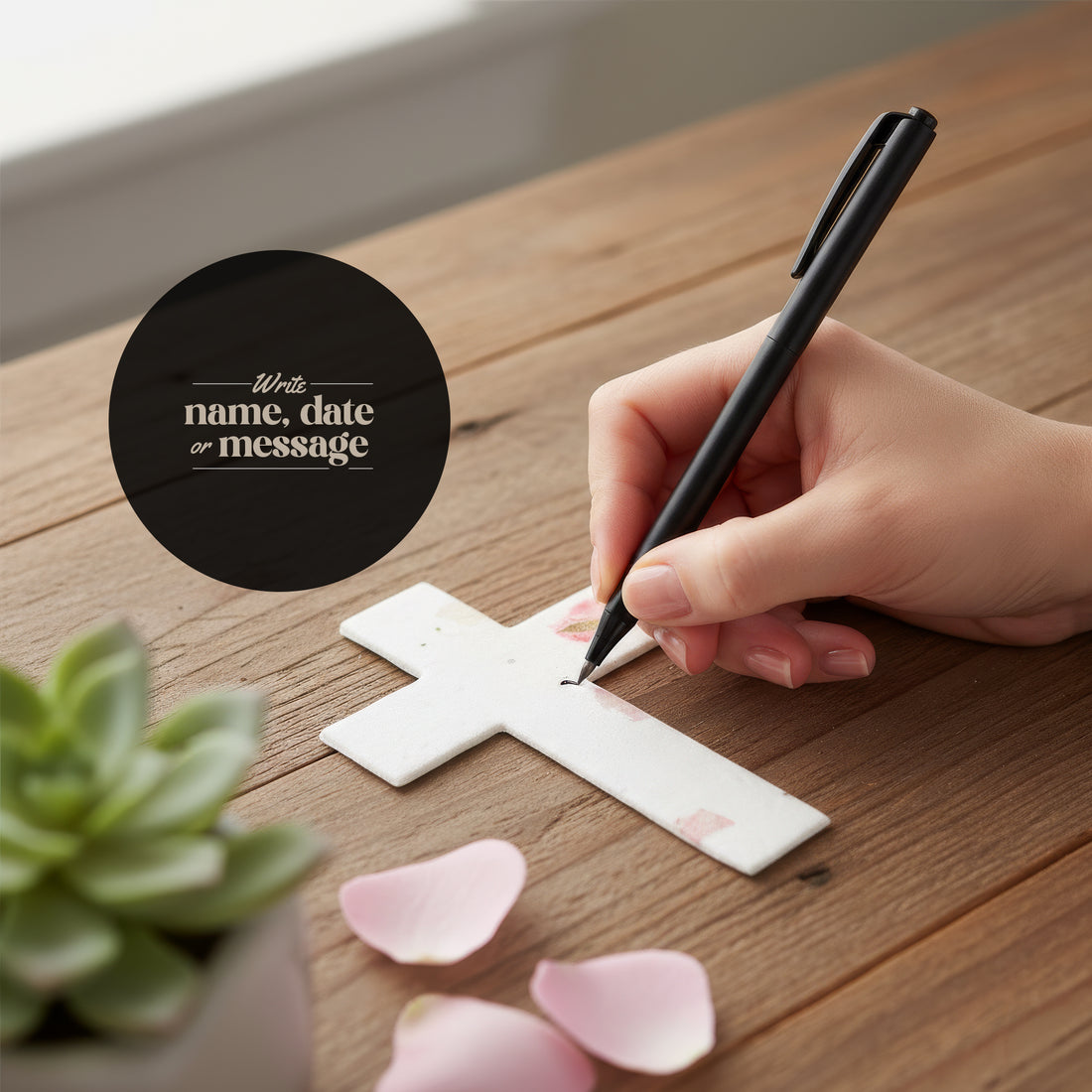 A hand writing on a white plantable seed paper cross funeral favor decorated with pressed pink flowers, using a black pen to add a name, date, or message on a wooden table