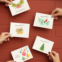 Five Plantable Holiday Christmas cards by Seed Bloom Gifts held by hands against a red brick wall.