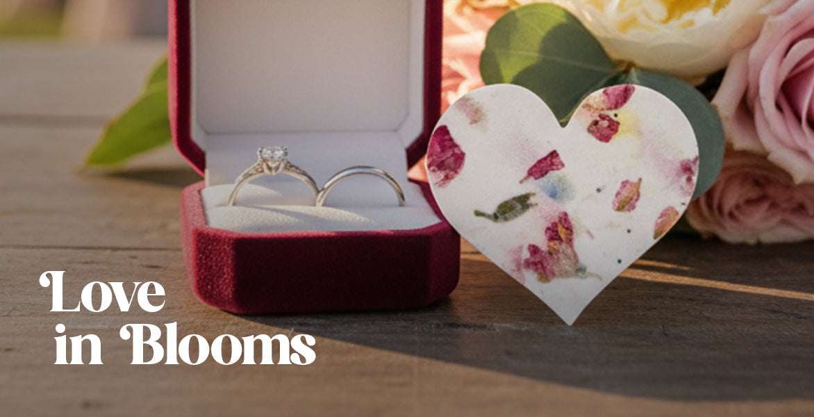 Two rings in a red box with a floral heart card and flowers on a wooden surface, text 'Love in Blooms'.