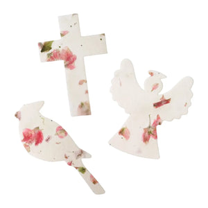 Three white bloom plantable seed paper funeral favor shapes decorated with pressed pink flowers and green leaves: a cross, a dove, and an angel figure