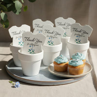 Plantable seed paper onesie baby shower favors arranged in white pots on a serving tray, displayed alongside blue-frosted cupcakes, each favor reading “Thank you for helping our baby bloom.”