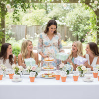 Outdoor baby shower scene with an expectant mother opening gifts surrounded by friends at a decorated table, featuring desserts, floral arrangements, and plantable seed paper onesie favors displayed in small orange terracotta pots.
