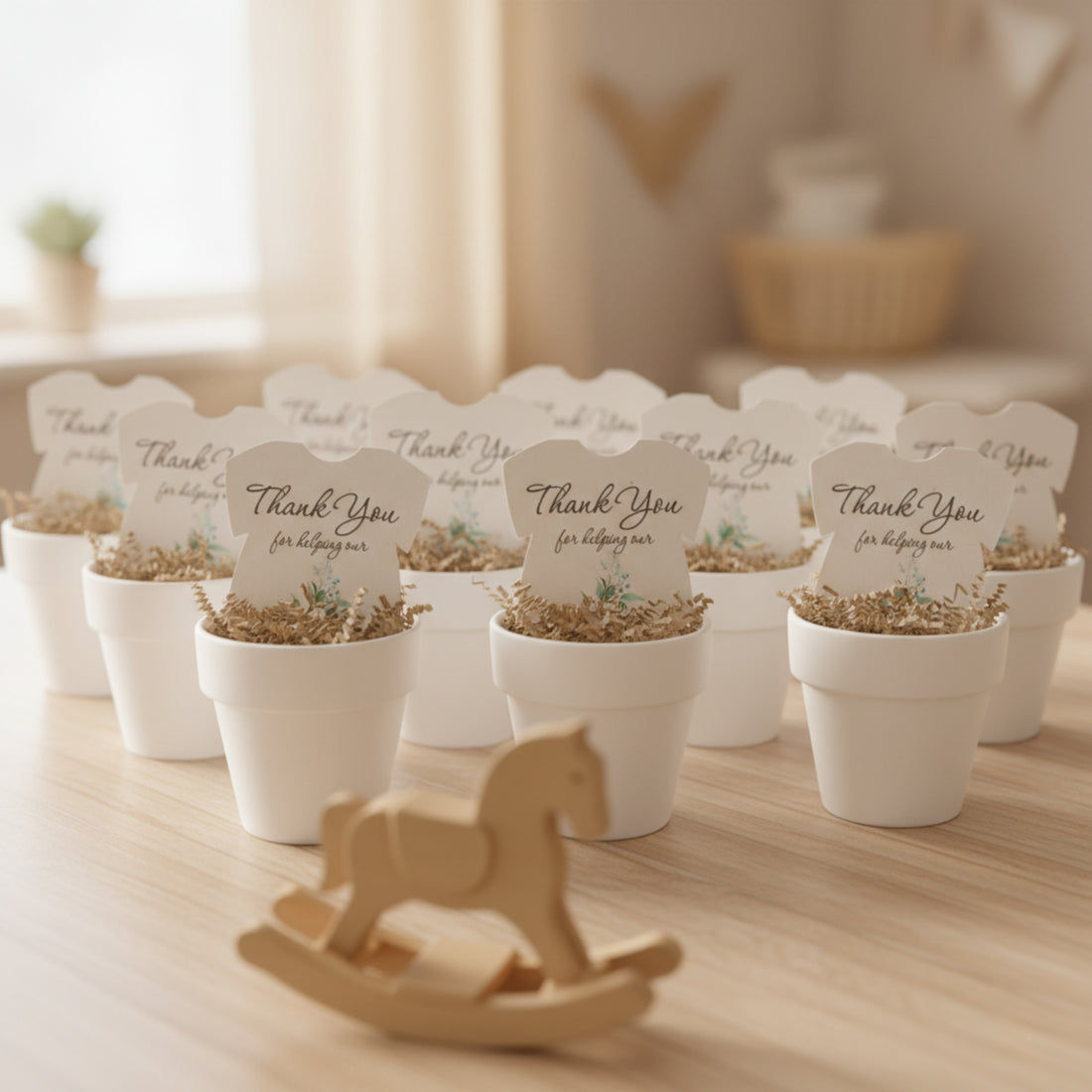 Plantable seed paper onesie baby shower favors arranged in white pots on a wooden table, featuring the message “Thank you for helping our baby bloom,” with soft nursery décor and a wooden rocking horse in the foreground.