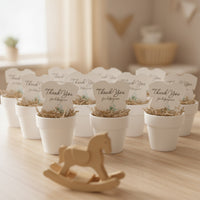 Plantable seed paper onesie baby shower favors arranged in white pots on a wooden table, featuring the message “Thank you for helping our baby bloom,” with soft nursery décor and a wooden rocking horse in the foreground.