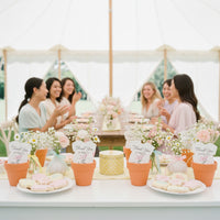 Baby shower table set with plantable seed paper onesie favors in orange terracotta pots, floral centerpieces, and desserts, with guests seated and chatting together under a bright outdoor tent.