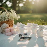 Outdoor baby shower picnic setup featuring plantable seed paper onesie favors in white pots, arranged on a blanket with a basket of flowers, baby booties, a plush toy, and ultrasound photos in soft sunlight.