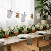 Baby shower table setup with plantable seed paper onesie favors in small terracotta pots at each place setting, decorated with greenery and a hanging garland of baby onesies and teddy bears.